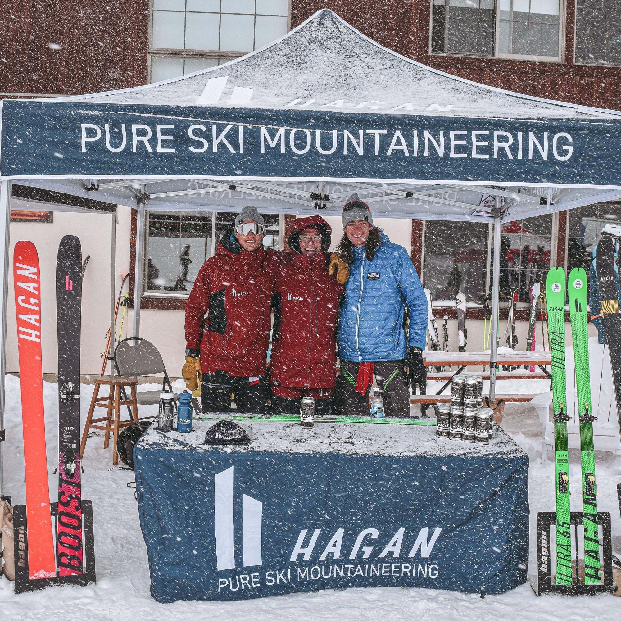 Three people standing under a 'Pure Ski Mountaineering' tent with snow falling.