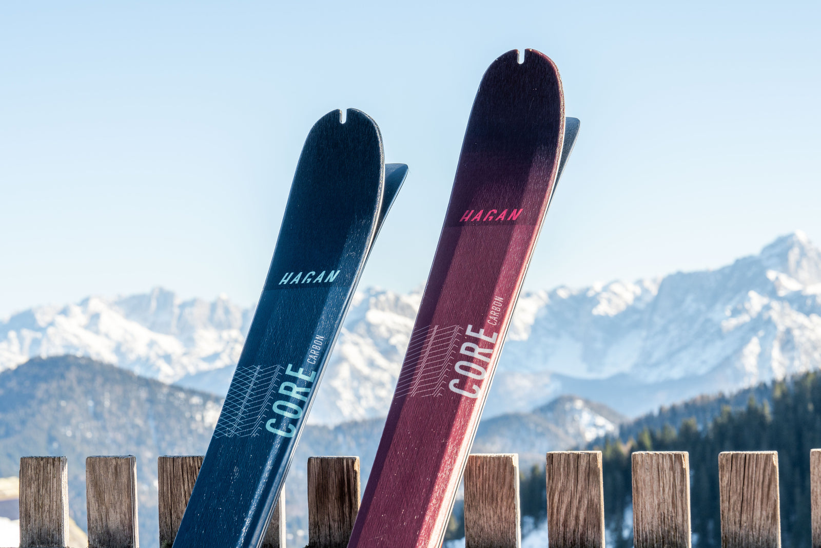 Two HAGAN Core Carbon skis lean against a fence with moutnains in the background.